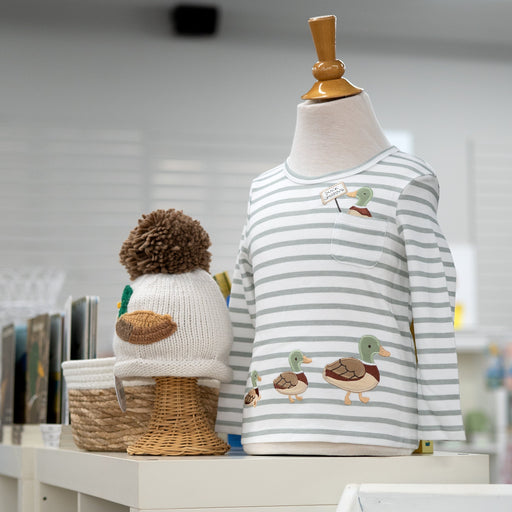 A children's long-sleeve striped shirt displayed on a mannequin torso, with duck illustrations on the front. A knitted duck hat sits beside it on a shelf in a softly lit indoor setting.