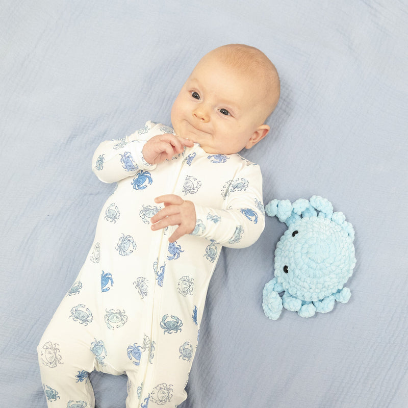 Baby in a white onesie with blue sea creature patterns lying on a light blue blanket next to a blue plush toy.