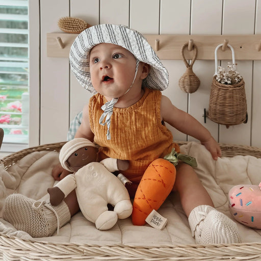 Baby in an orange outfit and striped hat sitting on a wicker bed with toys.