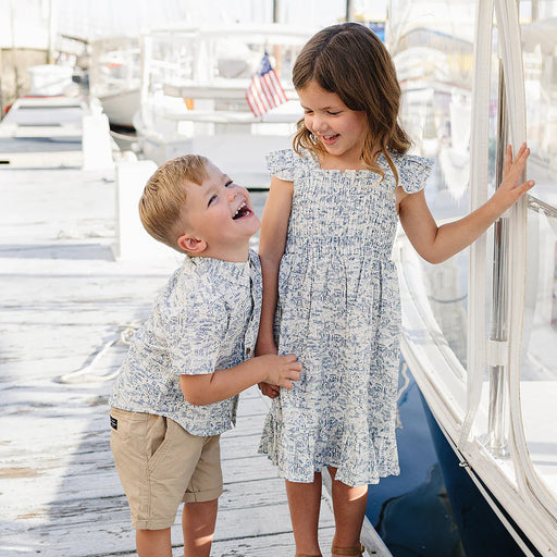 Two children, a boy and a girl, standing on a dock with boats in the background.