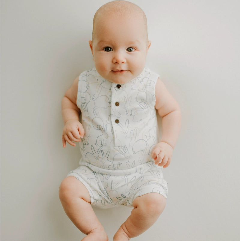 Baby wearing a white sleeveless romper with bunny patterns on a plain background