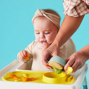 Baby in a high chair being fed with a spoon and tray against a blue background