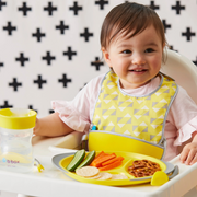 Child in a high chair with a yellow bib, smiling at the camera, with a plate of food and a sippy cup on the tray.