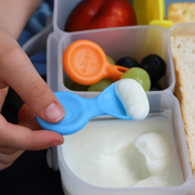Hand holding a blue spoon over a container of yogurt with a lunchbox in the background