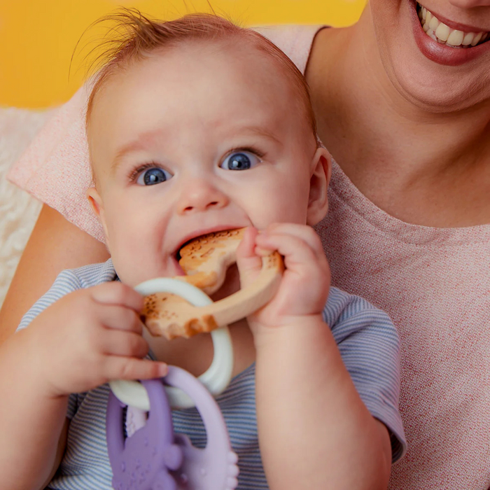 Baby holding a teething ring with a smiling adult in the background