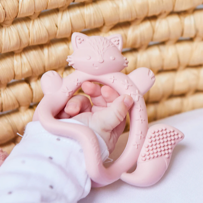 Pink fox-shaped teething ring held by a child's hand against a woven  basket background