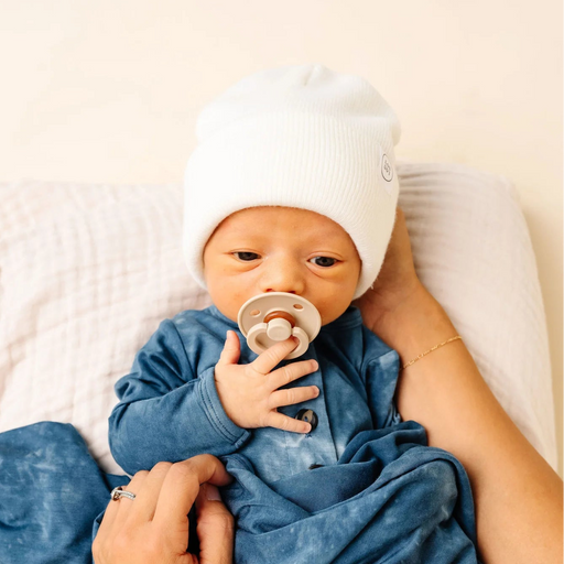 Newborn baby wrapped in blue blanket with ivory hat, holding a pacifier.
