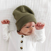 Baby wearing a moss green knit hat and white outfit with black buttons on a white background