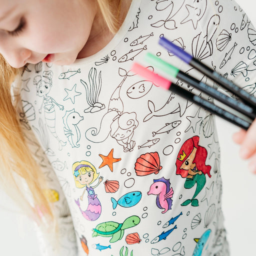 Child wearing Bee Colorful Co mermaid colorable pajamas while coloring the sea-themed design with fabric markers