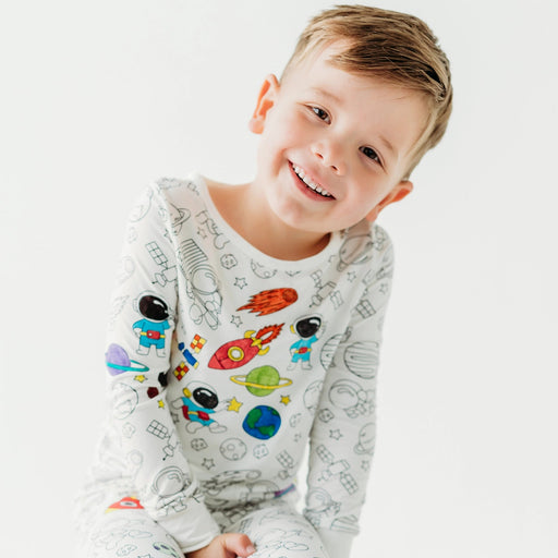 Child wearing Bee Colorful Co space colorable pajamas, smiling while seated on a stool against a white background