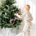Child in pajamas decorating a Christmas tree with red and gold ornaments on a white background