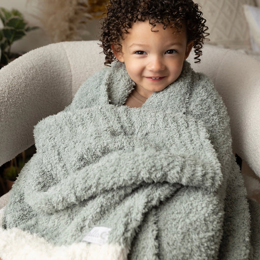 Child wrapped in sage Alani blanket, smiling in a chair