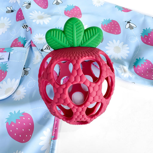Pink strawberry-shaped teething toy with green leaves on a blue fabric background with strawberry pattern
