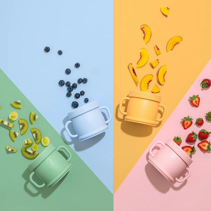 Three Coastal Cups in pink, blue, and sage green arranged on colorful pastel quadrants with snacks like blueberries, bananas, and strawberries scattered around.