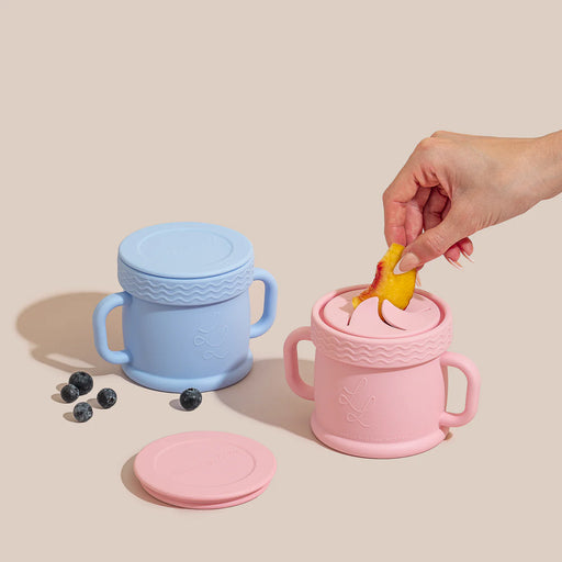 A pastel pink and blue Coastal Cup with lids shown on a neutral background, with a hand placing banana slices inside and scattered blueberries nearby.