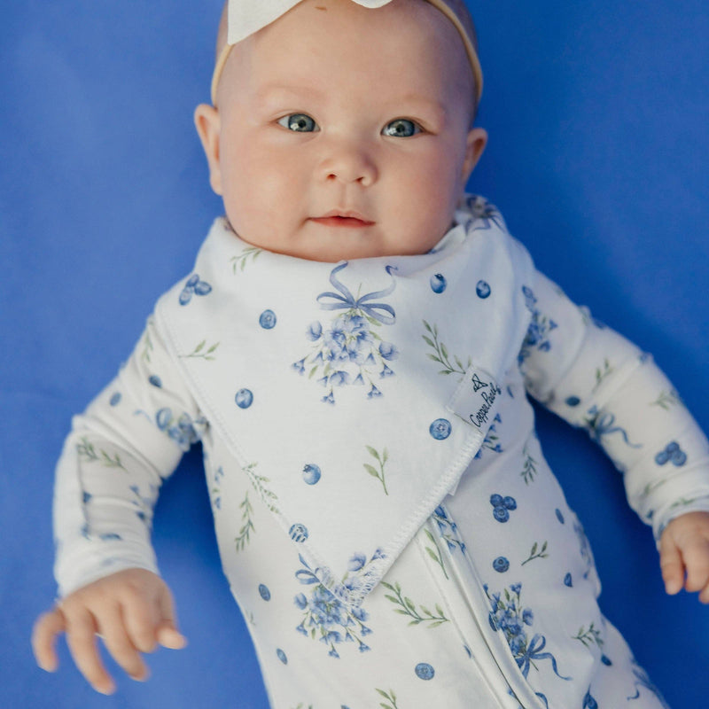 Baby lying on a blue background wearing the blue floral Copper Pearl Catherine bandana bib
