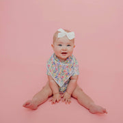 Baby sitting on a pink background wearing the wildflower Copper Pearl Catherine bandana bib