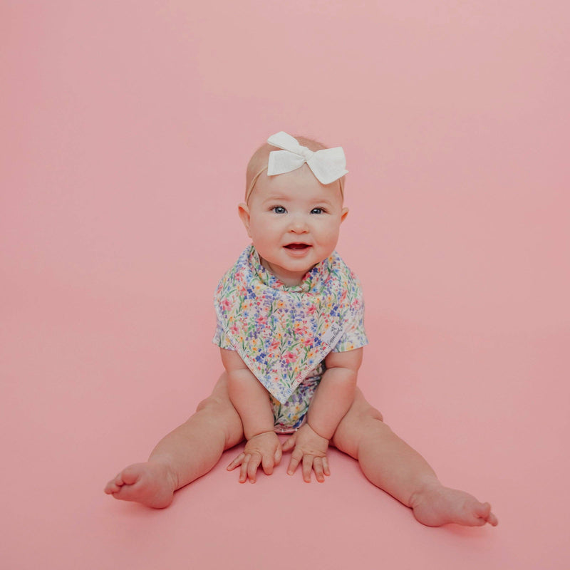 Baby sitting on a pink background wearing the wildflower Copper Pearl Catherine bandana bib