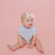 Baby sitting on a pink background wearing the blue gingham Copper Pearl Catherine bandana bib