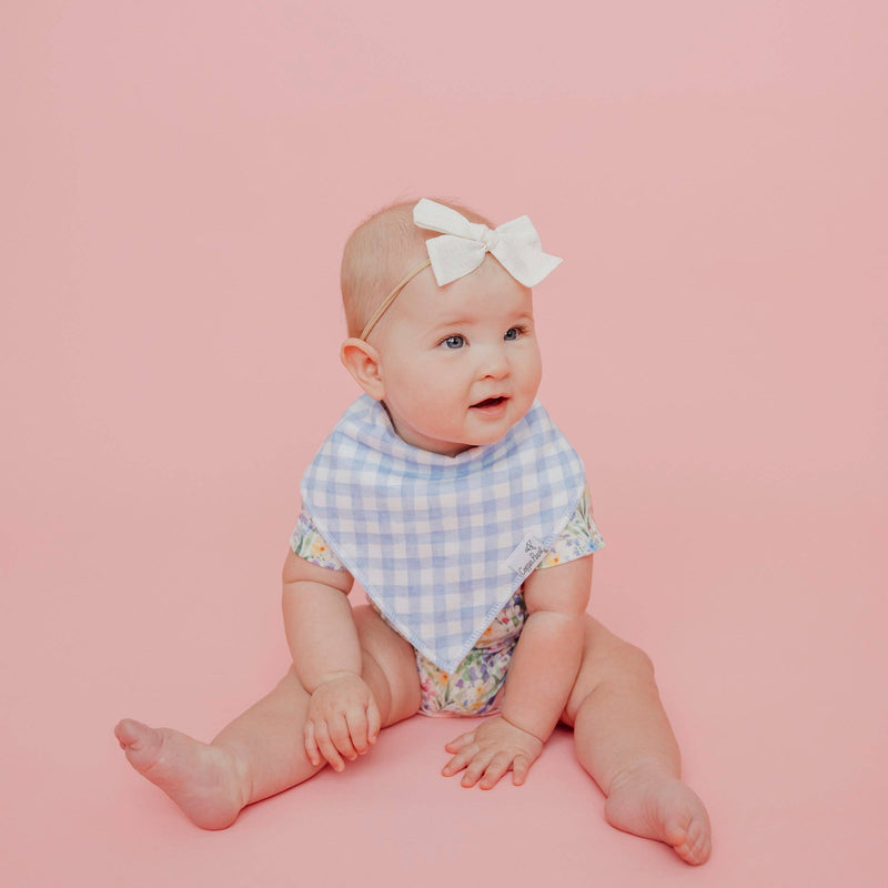 Baby sitting on a pink background wearing the blue gingham Copper Pearl Catherine bandana bib