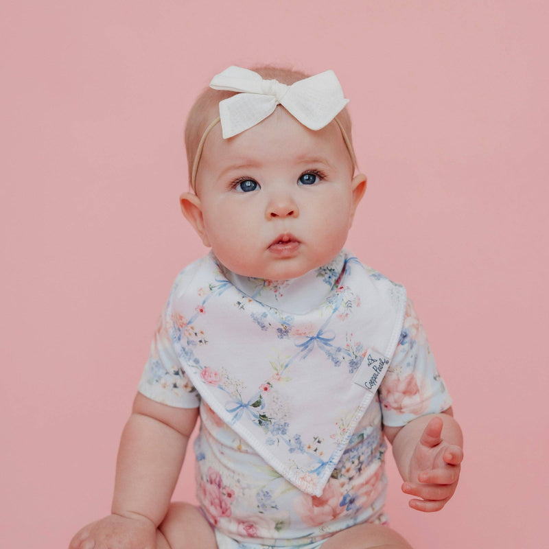 Baby with a white bow sitting on a pink background wearing a Copper Pearl Genevieve bandana bib