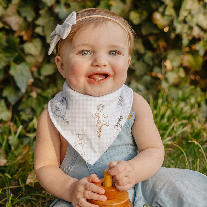 Smiling baby girl wearing a Copper Pearl Hundred Acre Wood Winnie the Pooh bandana bib outdoors