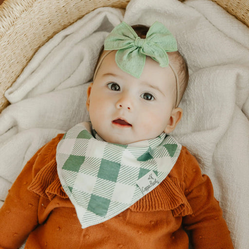 Baby wearing a green checkered bib and headband, lying on a soft surface.