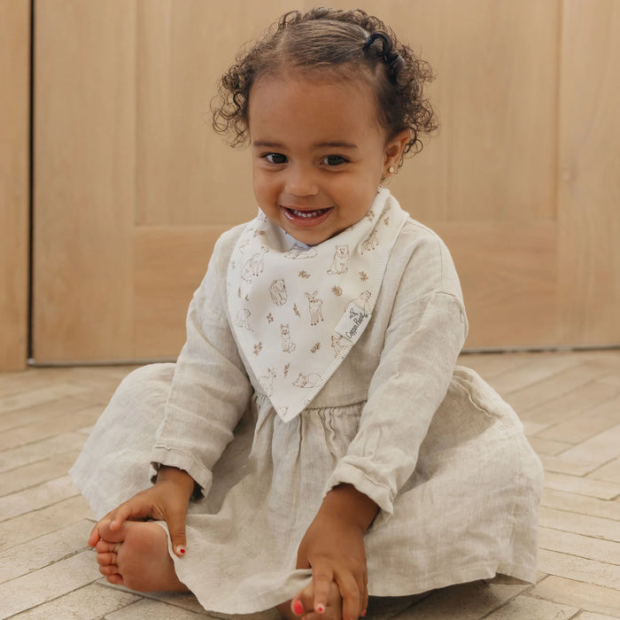Child wearing a light-colored dress and bib sitting on a wooden floor.