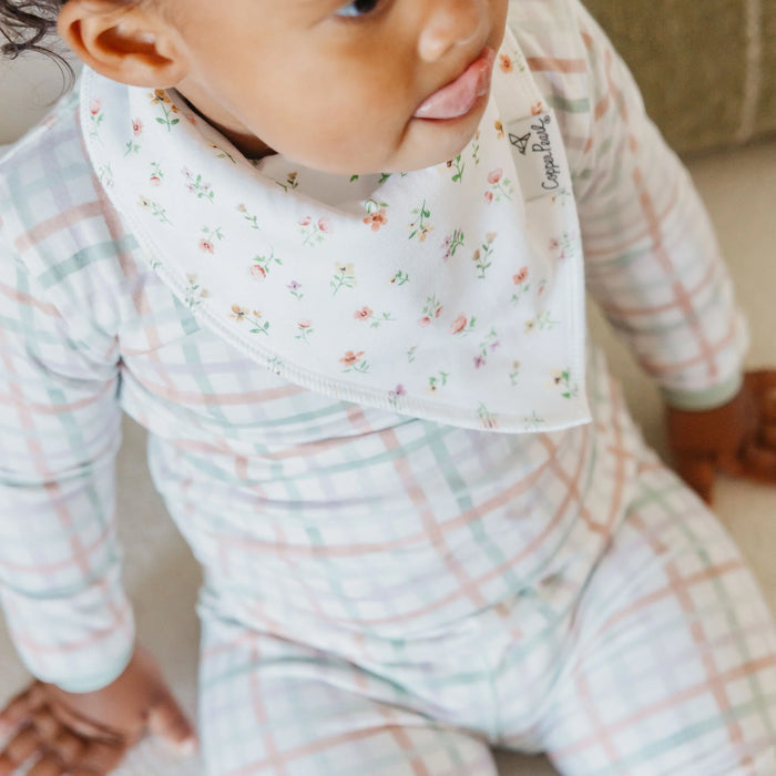 Baby wearing a floral bib and checkered outfit sitting on a couch.