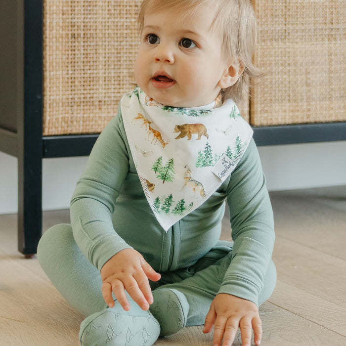 Baby wearing a green outfit and a bib with animal prints sitting on a wooden floor.