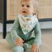 Baby wearing a green outfit and a bib with animal prints sitting on a wooden floor.
