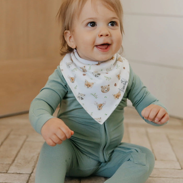 Baby wearing a green outfit and a bib with animal patterns on a wooden floor.