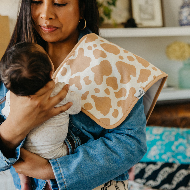 Parent holding baby with a Copper Pearl Boone burp cloth in tan cow print over the shoulder