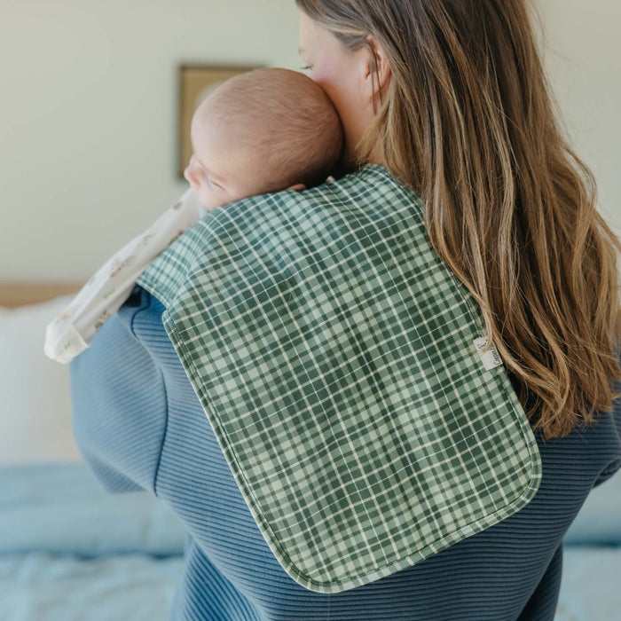 Woman holding a baby wrapped in a green checkered blanket in a bedroom setting.