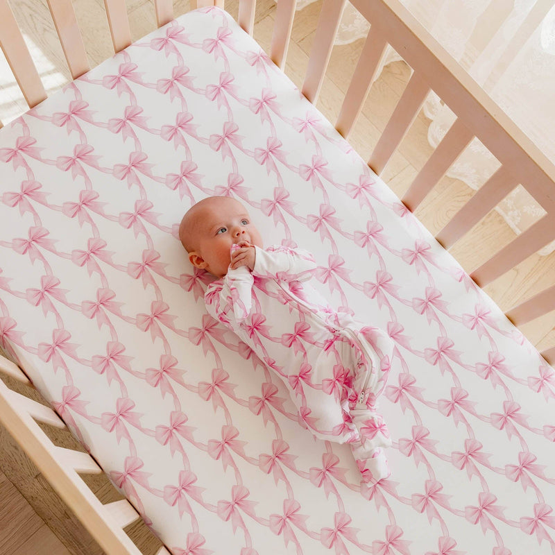 Angled view of a crib fitted with the Copper Pearl Bow My Gosh crib sheet with a baby lying on it