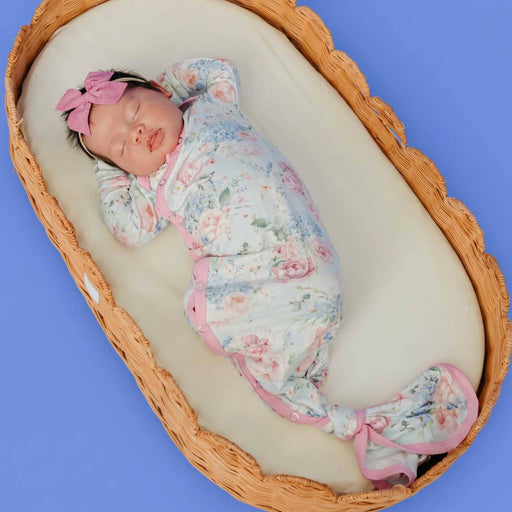 Newborn with a pink bow headband lying in a wicker basket wearing the Copper Pearl Genevieve knotted gown