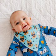 A baby lying on a white quilted blanket, wearing a blue long-sleeve romper and a matching light blue bib with a dog and police-themed print.