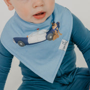 A baby wearing a light blue bib with a police car and dog graphic, paired with a blue long-sleeve top. The bib has a visible 'Lappin Baby' brand tag.