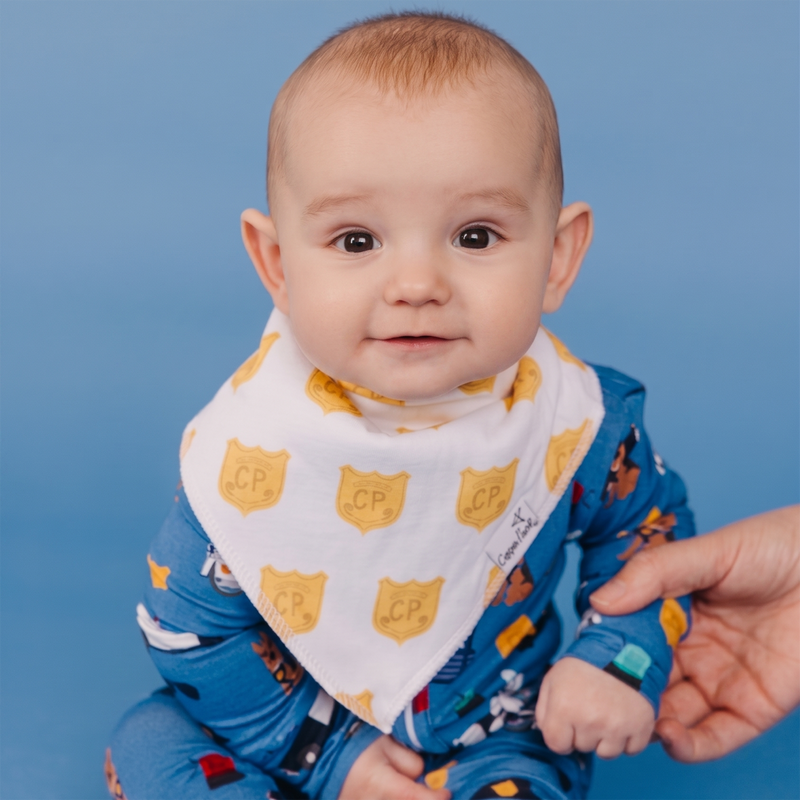 A baby wearing a white bandana bib with yellow shield patterns and a blue patterned onesie, held by an adult hand against a solid blue background.