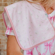 A close-up of a baby bib with a light pink background, white daisy floral print, and pink trim, worn by a person in a matching floral top.