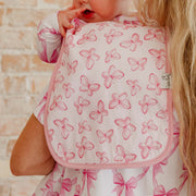 A close-up of a baby wearing a white bib with a pink butterfly pattern and pink trim, held by an adult. The background shows a light brick wall.