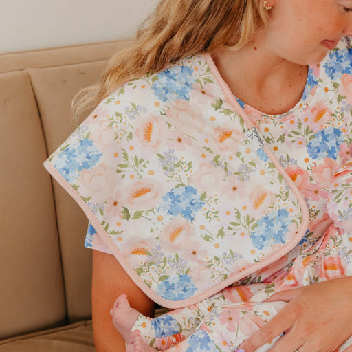 A close-up lifestyle shot of a woman wearing a floral print maternity nursing top and holding a baby. The top features a soft pink border and a delicate pattern of blue, pink, and orange flowers on a white background.
