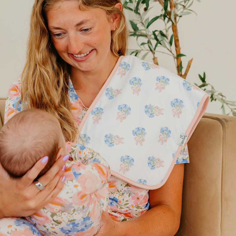 A woman in a floral robe holds a baby wearing a matching floral baby wrap or bib set. The fabric features a delicate pattern of blue flowers and pink leaves on a white background, with a solid pink border.
