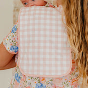 A close-up lifestyle shot of a baby wearing a pink and white gingham bib over a floral dress, held by a mother with long blonde hair.