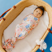 A baby swaddled in a floral-patterned blanket with pink, blue, and orange flowers on a white background, lying inside a woven Moses basket on a blue background.