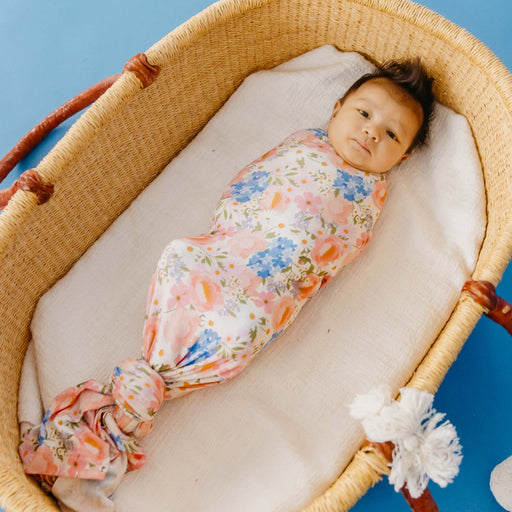 A baby swaddled in a floral-patterned blanket with pink, blue, and orange flowers on a white background, lying inside a woven Moses basket on a blue background.