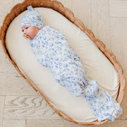 A baby swaddled in a light blue and white floral patterned swaddle blanket and matching bonnet, lying on a cream cushion inside a woven wicker bassinet on a light wood floor.