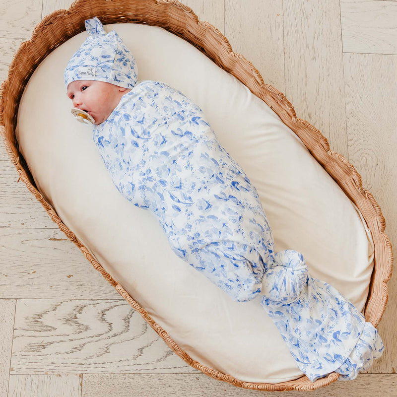 A baby swaddled in a light blue and white floral patterned swaddle blanket and matching bonnet, lying on a cream cushion inside a woven wicker bassinet on a light wood floor.
