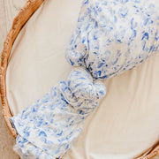 A close-up of a knotted baby swaddle blanket with a blue floral pattern on a white background, resting inside a woven wicker basket.
