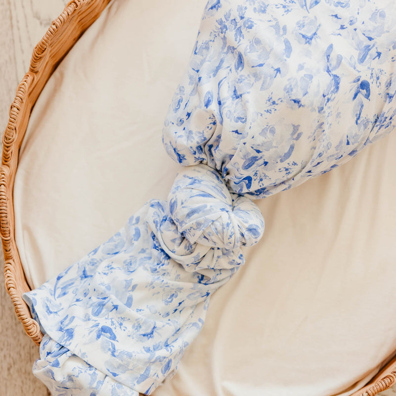 A close-up of a knotted baby swaddle blanket with a blue floral pattern on a white background, resting inside a woven wicker basket.